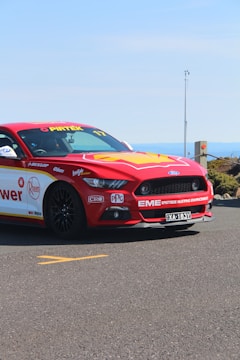 A red and yellow racing car, adorned with various sponsor logos, is parked on an asphalt road. It features a sleek design with sporty accents and a visible license plate. The background shows a clear blue sky and distant landscape.