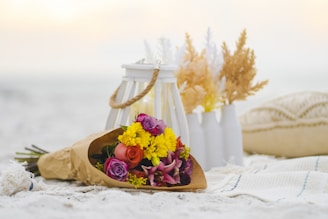 A bouquet of colorful flowers is placed on a sandy surface, wrapped in brown paper. Behind the bouquet, there is a white lantern with a rope handle and two white vases containing dried flowers. A textured cushion and a patterned cloth are also part of the setting, creating a serene and cozy ambiance.
