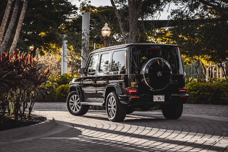A black luxury SUV with a Mercedes-Benz logo, parked on a cobblestone driveway surrounded by lush greenery and tropical plants. The scene is set during the late afternoon with sunlight creating a warm glow.