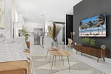 A modern living room with a minimalist design features a brown sofa with white pillows, a striped rug, and nested wooden coffee tables. There are decorative plants and a large black accent wall displaying a flat-screen TV showing a beach scene. The room is well-lit and extends into an open kitchen area with white cabinetry and stainless steel appliances.