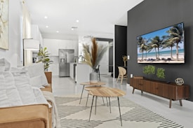 A modern living room with a minimalist design features a brown sofa with white pillows, a striped rug, and nested wooden coffee tables. There are decorative plants and a large black accent wall displaying a flat-screen TV showing a beach scene. The room is well-lit and extends into an open kitchen area with white cabinetry and stainless steel appliances.