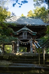 A traditional wooden temple gate framed by greenery, with steps leading up to it. The structure features intricate carvings and a rope with paper tassels hanging across the entrance. Sunlight filters through the trees, casting shadows on the path and building.