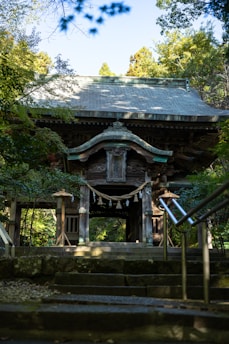 A traditional wooden temple gate framed by greenery, with steps leading up to it. The structure features intricate carvings and a rope with paper tassels hanging across the entrance. Sunlight filters through the trees, casting shadows on the path and building.