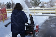 A person in a dark blue jacket is operating a snowblower to clear a walkway covered in snow. The snow is being thrown to the side, forming a spray in the air. There is a yellow railing and a red safety barrier nearby. Snow is covering the ground and trees in the background.