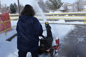 A person in a dark blue jacket is operating a snowblower to clear a walkway covered in snow. The snow is being thrown to the side, forming a spray in the air. There is a yellow railing and a red safety barrier nearby. Snow is covering the ground and trees in the background.