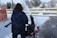 A person in a dark blue jacket is operating a snowblower to clear a walkway covered in snow. The snow is being thrown to the side, forming a spray in the air. There is a yellow railing and a red safety barrier nearby. Snow is covering the ground and trees in the background.
