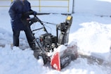 A person is using a snowblower to clear snow from a paved surface. The surrounding area is covered in fresh, white snow, and there's a yellow rail and chain-link fence in the background. The person is wearing a dark blue coat and jeans, and the snowblower has a visible red section.