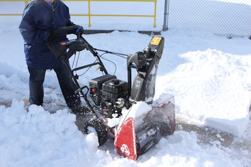 A person is using a snowblower to clear snow from a paved surface. The surrounding area is covered in fresh, white snow, and there's a yellow rail and chain-link fence in the background. The person is wearing a dark blue coat and jeans, and the snowblower has a visible red section.