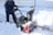 A person is using a snowblower to clear snow from a paved surface. The surrounding area is covered in fresh, white snow, and there's a yellow rail and chain-link fence in the background. The person is wearing a dark blue coat and jeans, and the snowblower has a visible red section.