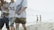 Several people are walking on a sandy beach near the shoreline, with some wearing casual summer attire. Two individuals are seen in the background wading in the water.