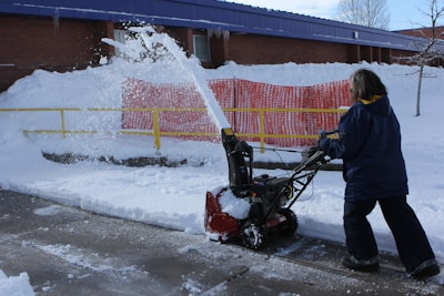 A person is operating a snow blower, clearing snow from a sidewalk. Snow is being discharged in an arc through the air. A bright yellow railing and a red safety barrier are visible in the background, lining the snow-covered embankment alongside a brick building.