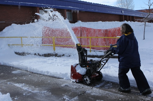 A person is operating a snow blower, clearing snow from a sidewalk. Snow is being discharged in an arc through the air. A bright yellow railing and a red safety barrier are visible in the background, lining the snow-covered embankment alongside a brick building.