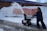 A person is operating a snow blower, clearing snow from a sidewalk. Snow is being discharged in an arc through the air. A bright yellow railing and a red safety barrier are visible in the background, lining the snow-covered embankment alongside a brick building.