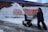 A person is operating a snow blower, clearing snow from a sidewalk. Snow is being discharged in an arc through the air. A bright yellow railing and a red safety barrier are visible in the background, lining the snow-covered embankment alongside a brick building.