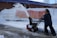 A person is operating a snow blower, clearing snow from a sidewalk. Snow is being discharged in an arc through the air. A bright yellow railing and a red safety barrier are visible in the background, lining the snow-covered embankment alongside a brick building.
