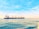 A large cargo ship with multiple cranes on deck navigates through calm ocean waters under a bright, clear sky. The ship has a blue and white color scheme with the word 'SEROS' visible on the side. Sunlight reflects off the water's surface, adding golden hues to the scene.