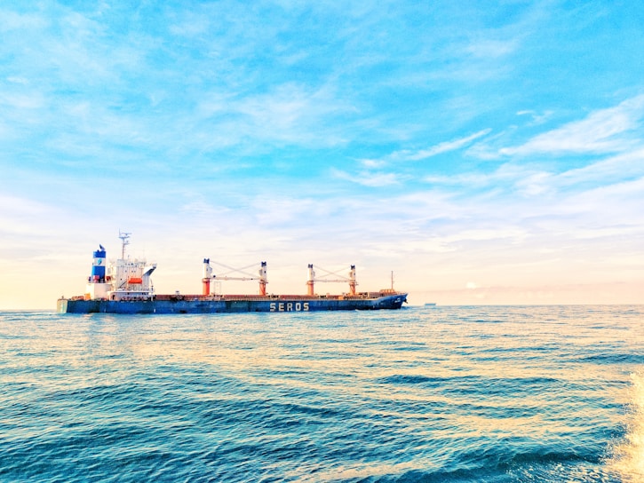 A large cargo ship with multiple cranes on deck navigates through calm ocean waters under a bright, clear sky. The ship has a blue and white color scheme with the word 'SEROS' visible on the side. Sunlight reflects off the water's surface, adding golden hues to the scene.