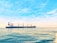 A large cargo ship with multiple cranes on deck navigates through calm ocean waters under a bright, clear sky. The ship has a blue and white color scheme with the word 'SEROS' visible on the side. Sunlight reflects off the water's surface, adding golden hues to the scene.