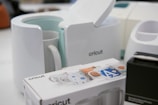 A Cricut mug press is displayed alongside a Cricut box featuring decorative designs for mugs, as well as other devices in the background. The scene seems to be set in a workspace or creative area.