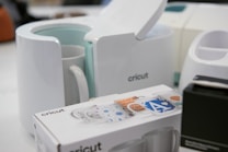 A Cricut mug press is displayed alongside a Cricut box featuring decorative designs for mugs, as well as other devices in the background. The scene seems to be set in a workspace or creative area.