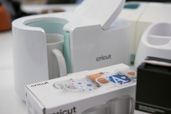 A Cricut mug press is displayed alongside a Cricut box featuring decorative designs for mugs, as well as other devices in the background. The scene seems to be set in a workspace or creative area.