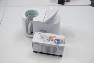 A Cricut mug press with its lid open is placed on a table alongside a box containing two mugs. The box displays colorful mug designs. The setting is a plain white surface, creating a clean and minimalist appearance.