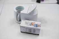 A Cricut mug press with its lid open is placed on a table alongside a box containing two mugs. The box displays colorful mug designs. The setting is a plain white surface, creating a clean and minimalist appearance.