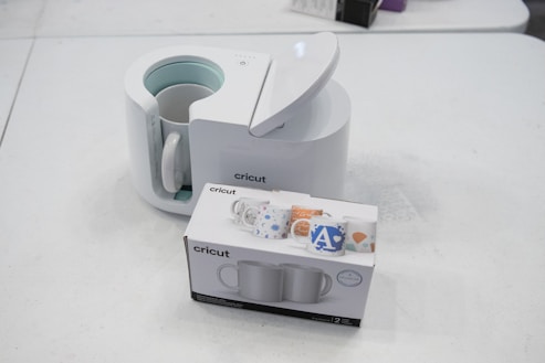 A Cricut mug press with its lid open is placed on a table alongside a box containing two mugs. The box displays colorful mug designs. The setting is a plain white surface, creating a clean and minimalist appearance.