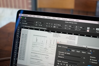 A close-up of a computer screen displaying Adobe InDesign 2023 software. The screen shows a detailed layout of a document with various text, tables, and boxes. The toolbars and text formatting options are visible on the interface. In the background, there's a wooden table and a blurred out-of-focus area.