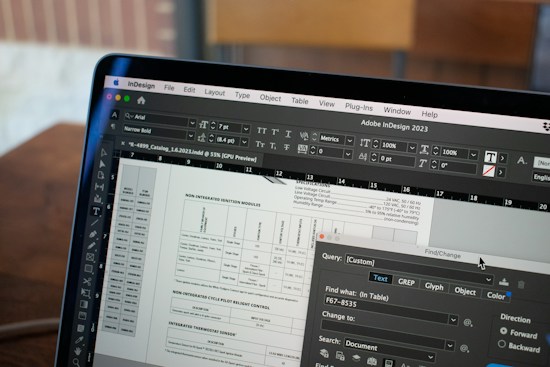 A close-up of a computer screen displaying Adobe InDesign 2023 software. The screen shows a detailed layout of a document with various text, tables, and boxes. The toolbars and text formatting options are visible on the interface. In the background, there's a wooden table and a blurred out-of-focus area.