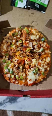 A small pizza topped with cheese, bell peppers, onions, and red chili flakes is placed inside an open cardboard box. Two small green and black sauce packets are visible in the background.