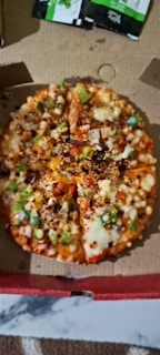 A small pizza topped with cheese, bell peppers, onions, and red chili flakes is placed inside an open cardboard box. Two small green and black sauce packets are visible in the background.