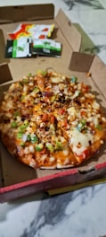 A pizza topped with a variety of ingredients such as green peppers, onions, cheese, and sprinkled spices is placed in an open cardboard pizza box. There are sauce packets visible in the background.