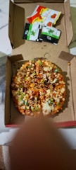 A pizza with various toppings, including vegetables and perhaps meat, is inside a partially open cardboard box. The pizza is cut into several slices. Next to the pizza, there are four condiment packets, one red, one yellow, and two black and green.