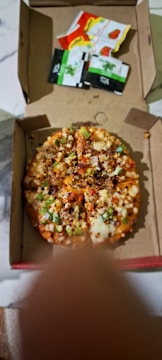 A pizza with various toppings, including vegetables and perhaps meat, is inside a partially open cardboard box. The pizza is cut into several slices. Next to the pizza, there are four condiment packets, one red, one yellow, and two black and green.