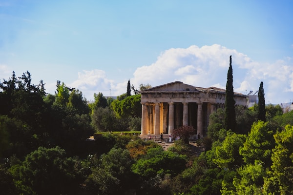 An ancient Greek temple with classical columns is surrounded by lush greenery and trees under a partly cloudy sky. The temple is slightly illuminated by sunlight, creating a warm contrast against the green landscape.