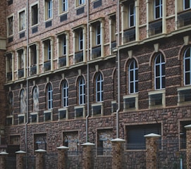 A facade of a building featuring multiple stories with repeated patterns of arched windows and brickwork. Dark brown bricks form the dominant design, with lighter bricks highlighting the window frames. The lower part of the structure includes a black metal fence with brick columns.