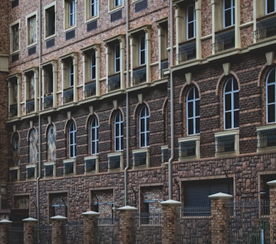 A facade of a building featuring multiple stories with repeated patterns of arched windows and brickwork. Dark brown bricks form the dominant design, with lighter bricks highlighting the window frames. The lower part of the structure includes a black metal fence with brick columns.