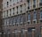 A facade of a building featuring multiple stories with repeated patterns of arched windows and brickwork. Dark brown bricks form the dominant design, with lighter bricks highlighting the window frames. The lower part of the structure includes a black metal fence with brick columns.