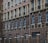 A facade of a building featuring multiple stories with repeated patterns of arched windows and brickwork. Dark brown bricks form the dominant design, with lighter bricks highlighting the window frames. The lower part of the structure includes a black metal fence with brick columns.