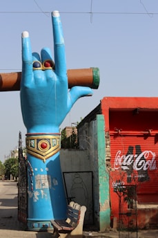A large, blue sculpture of a hand making a peace sign, adorned with rings and decorative elements, stands beside a red building with a Coca-Cola sign. The background includes a light blue wall and a metal gate. A wooden bench is positioned nearby.
