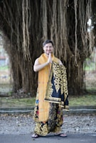 A person stands outdoors dressed in ornate traditional clothing with detailed patterns, combining bright orange and black with gold designs. The background consists of large, hanging tree roots or vines, creating a natural and serene setting.