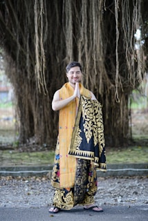 A person stands outdoors dressed in ornate traditional clothing with detailed patterns, combining bright orange and black with gold designs. The background consists of large, hanging tree roots or vines, creating a natural and serene setting.