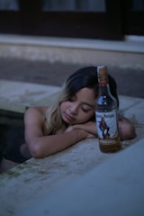 A person with closed eyes leans on a surface next to a bottle of Captain Morgan spiced rum. The setting appears to be relaxed with a subdued lighting environment, possibly by a pool.