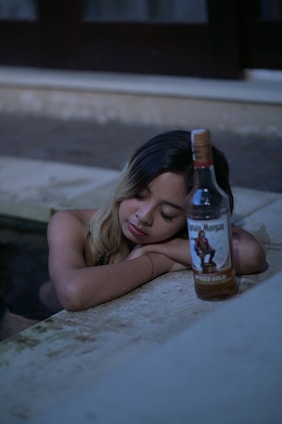 A person with closed eyes leans on a surface next to a bottle of Captain Morgan spiced rum. The setting appears to be relaxed with a subdued lighting environment, possibly by a pool.