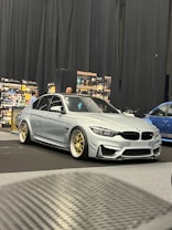 A silver BMW sports car is parked inside a showroom with a wooden floor and surrounded by other vehicles. It features gold alloy wheels and is positioned next to shelves stocked with various bottles, suggesting it might be part of an exhibition or trade show. A person is visible in the background.