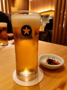 A tall glass of beer with a frothy head on a wooden table, featuring a Sapporo logo. A small dish with a garnished appetizer is placed next to the glass. Soft, warm lighting suggests a cozy interior, with blurred figures in the background, likely in a restaurant setting.