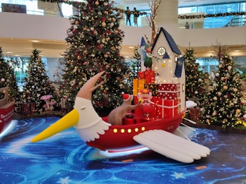 A festive Christmas scene with decorated trees and a whimsical flying ship. The ship has a bird-like design with a long yellow beak and a red body. It carries a house stacked with red and gold presents. A fairy holding a star is perched near the top, and a pink character with antlers sits at the ship’s base. The background shows a shopping mall interior adorned with garlands and lights.