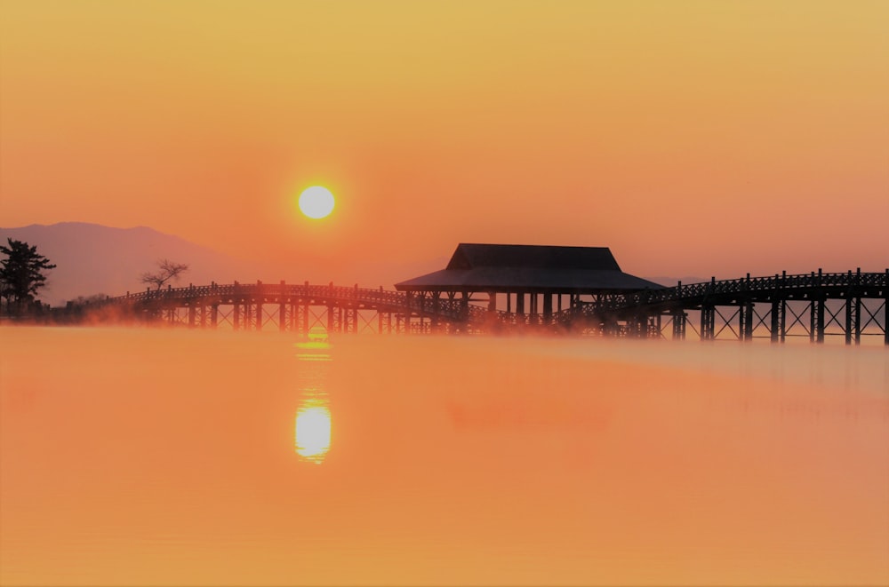 the sun is setting over the water with a pier in the background