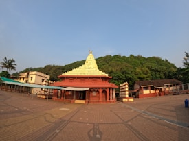 A traditional temple with a red and gold gopuram stands in an open courtyard. The temple structure features ornate carvings and is framed by lush green trees in the background. Nearby, several smaller buildings and a row of shaded canopies are visible. The area is paved with stone tiles, and the sky is clear with a soft blue hue.